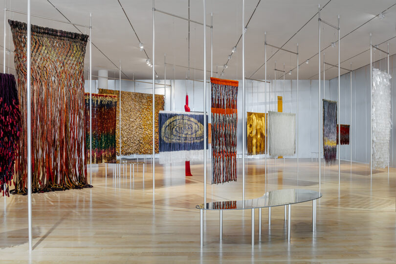A modern gallery space displays various large, colorful woven textile artworks hanging from the ceiling, with a reflective bench on the wooden floor.