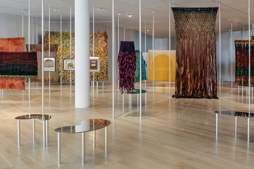 Contemporary art gallery with colorful textile installations hanging from the ceiling and mirrored benches on a light wooden floor.