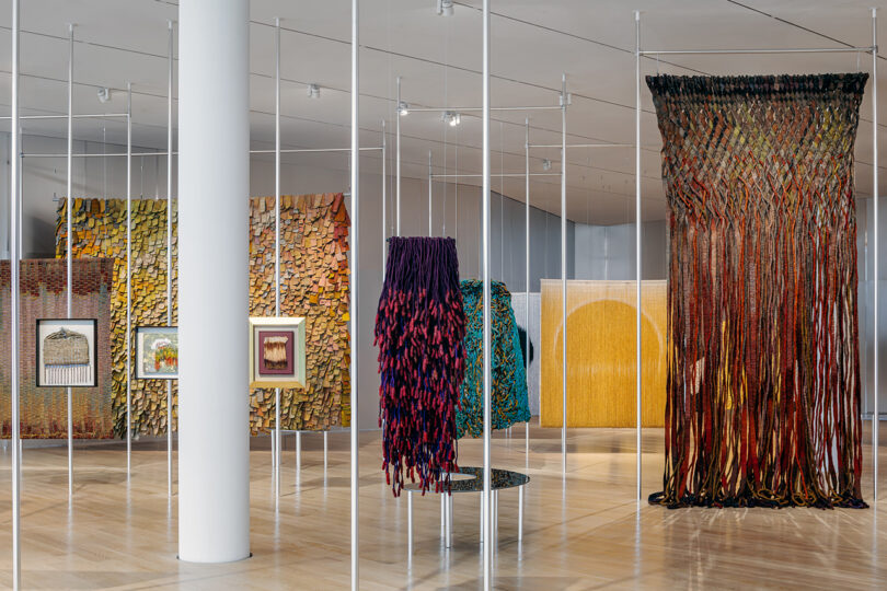 Art installation featuring textile and woven works, framed pieces, and wall-mounted sculptures displayed in a spacious, modern gallery with polished floors and white walls.