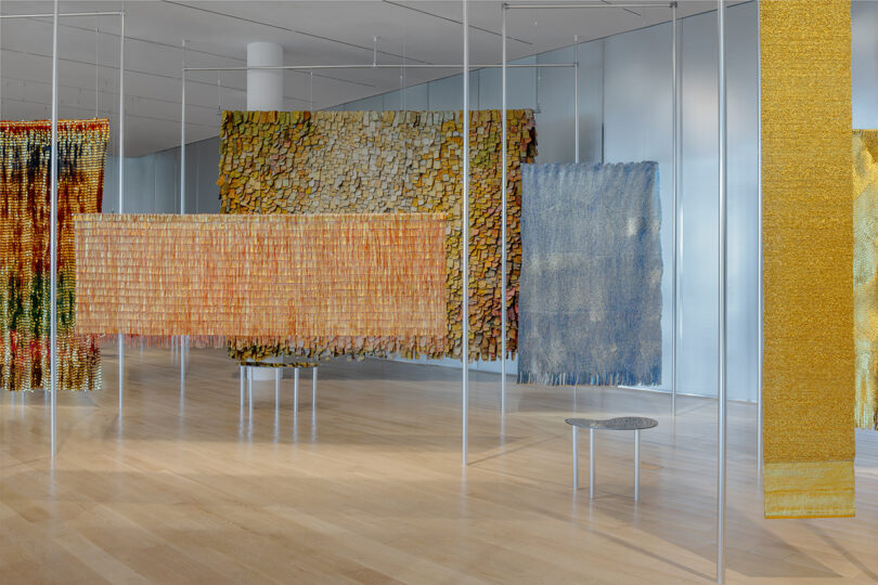 Multiple large woven textile artworks hang from the ceiling in a modern gallery space with wooden floors, white walls, and minimal furnishings.