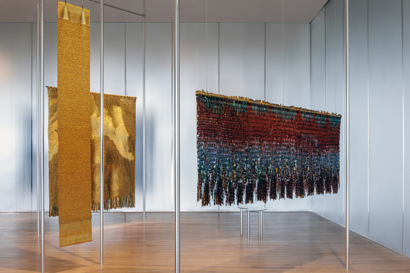 Two large woven textile artworks, one gold and one multicolored, hang vertically from ceiling-mounted supports in a modern gallery with wood floors and gray walls.