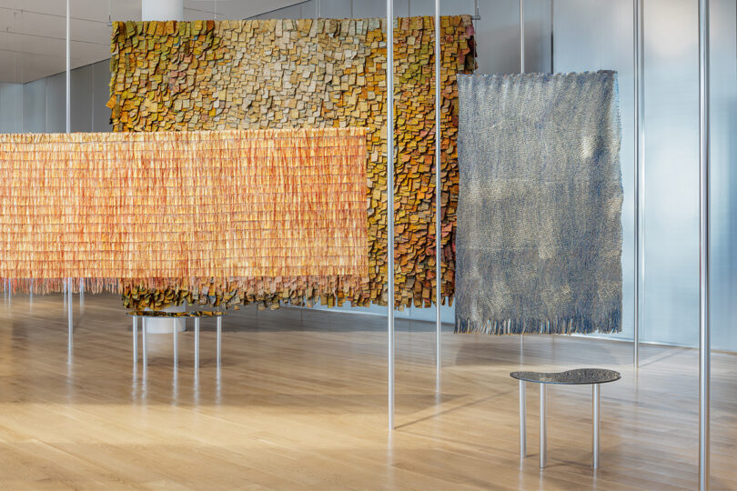 Three textured hanging textile artworks in shades of yellow, orange, and gray are displayed in a modern gallery with wooden floors and a small bench.
