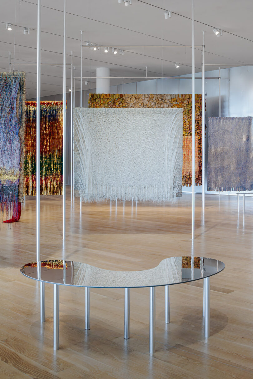A reflective silver bench sits on a wooden floor in front of several large, hanging textile artworks in a modern gallery space.