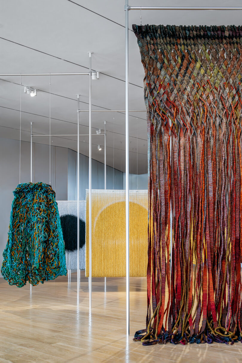 Installation view of large, colorful woven and knotted textile artworks hanging in a modern gallery space with light wood floors and white walls.