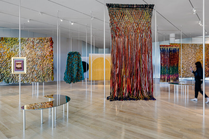 Large contemporary art gallery with colorful textile installations hanging from ceiling and walls, mirrored tables, and a person walking through the space.
