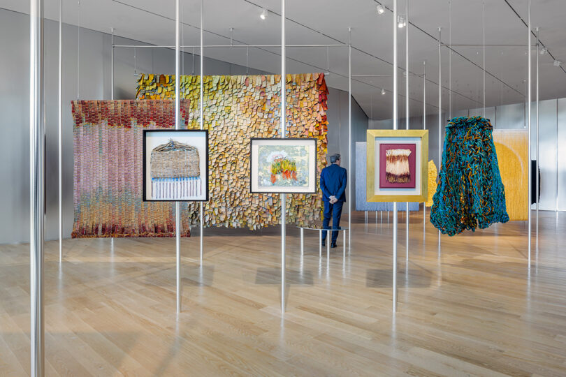A person stands in a modern art gallery viewing colorful, textured textile artworks and framed pieces hanging from the ceiling on display.