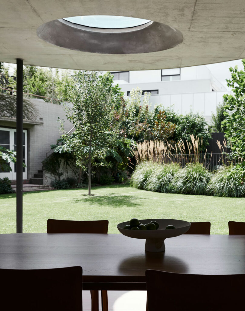 A dining table with a bowl of fruit sits indoors beneath a circular skylight, overlooking a modern backyard garden with trees and shrubs.
