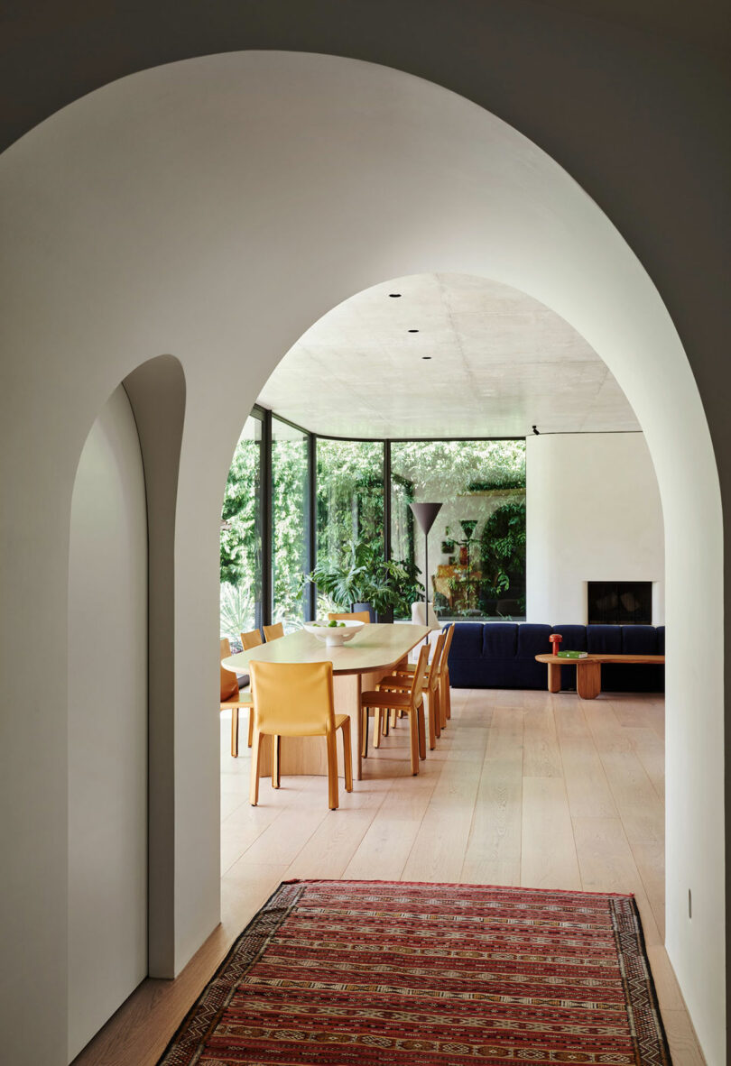 Modern dining room with yellow chairs and a wooden table, viewed through a white archway. Large windows overlook greenery; a dark sofa and red rug are also visible.