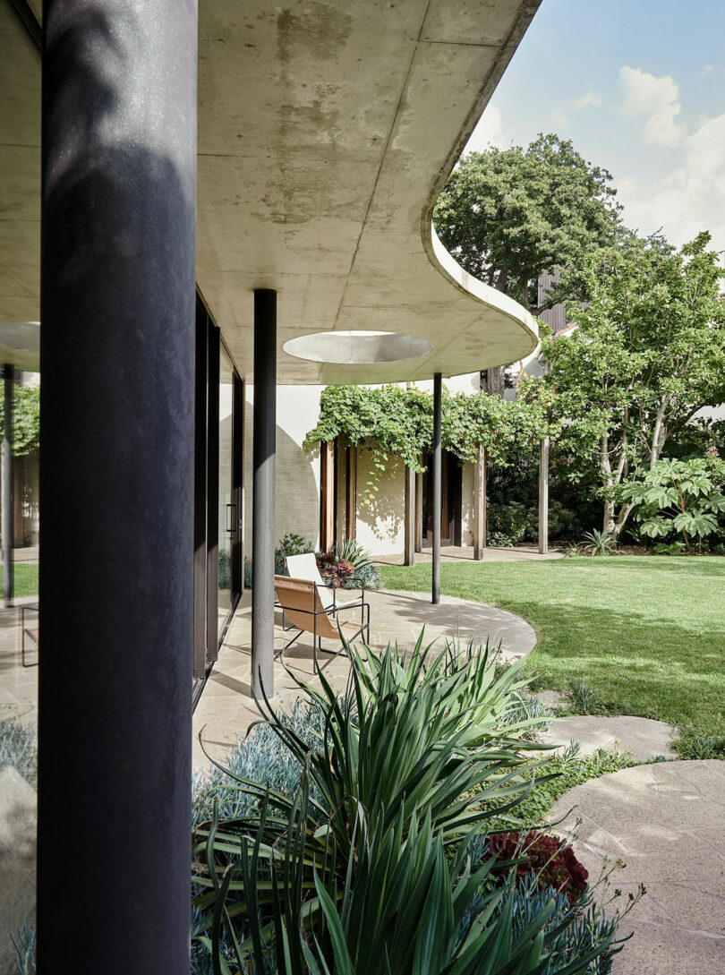 Modern house with curved concrete roof, black columns, glass walls, and a landscaped garden with greenery and outdoor seating.