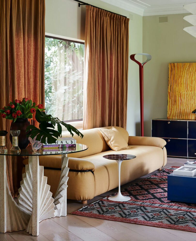 A living room with a mustard yellow sofa, geometric glass table, patterned rug, floor lamp, books, and wall art; large window with rust-colored curtains.