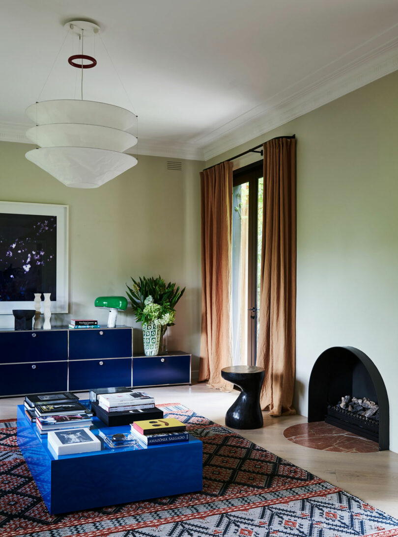 Modern living room with a blue sideboard, blue coffee table, books, patterned rug, fireplace, large window with tan curtains, and a white pendant light fixture.