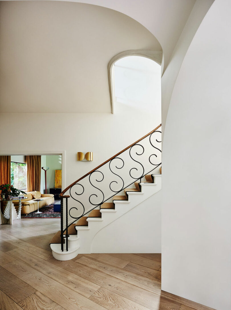 A modern interior with a wooden staircase featuring a decorative black railing, light wood floors, and an arched doorway leading to a living room with gold curtains.