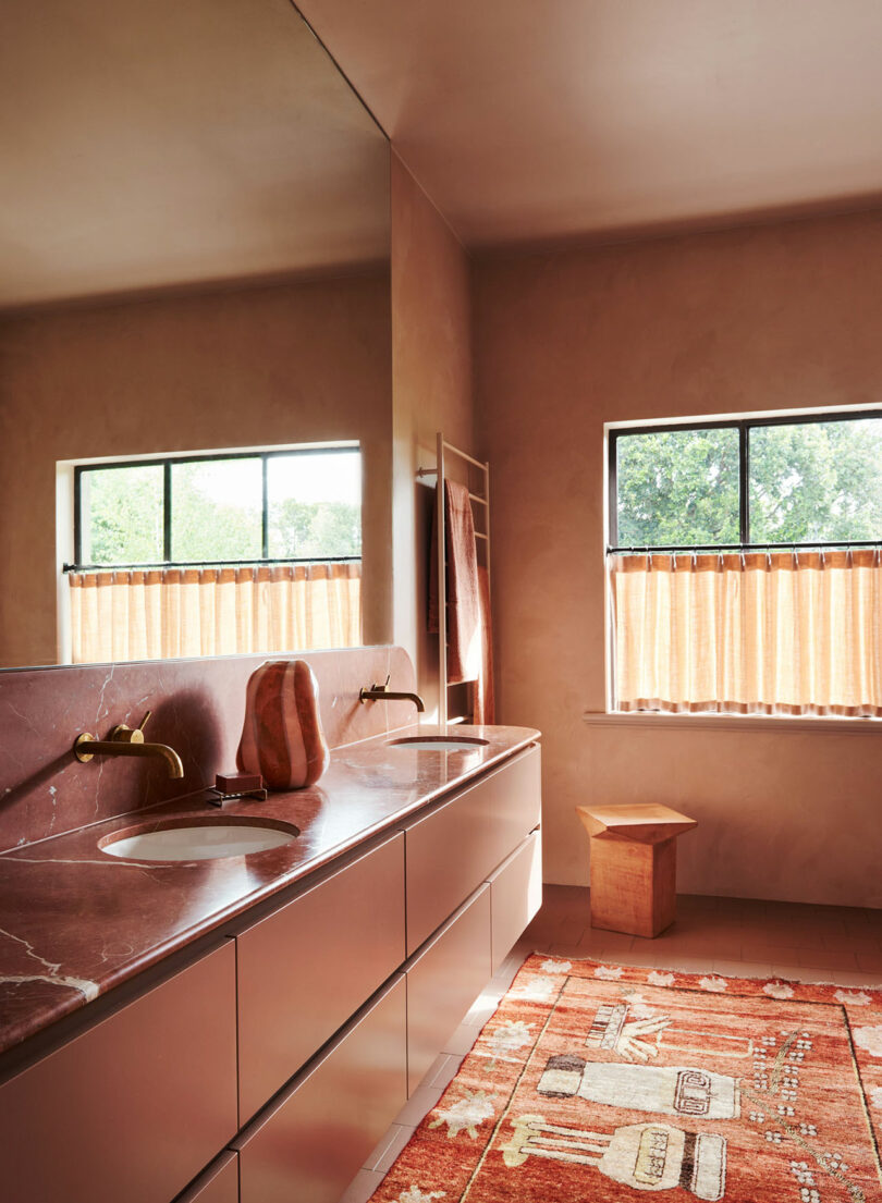Modern bathroom with a long marble countertop, double sinks, a large mirror, window with peach curtains, wooden stool, and patterned orange rug on the floor.