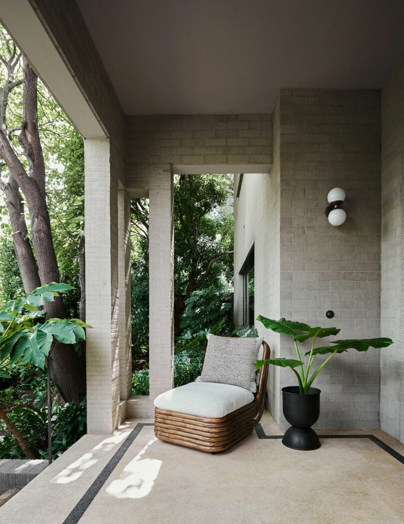 A wicker lounge chair with a cushion sits on a covered patio with beige brick walls, next to a black planter with large green leaves and surrounded by trees and greenery.