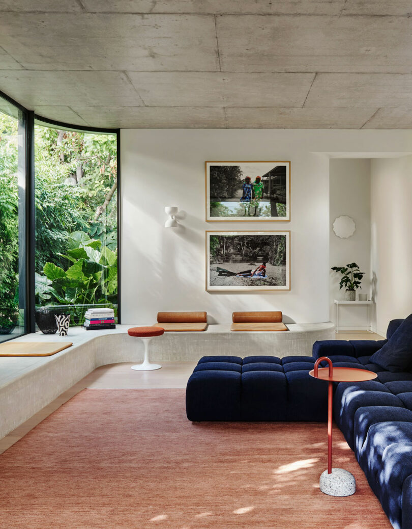 A modern living room with large windows, navy modular sofa, orange rug, two wall art pieces, indoor plants, and a small round white side table.