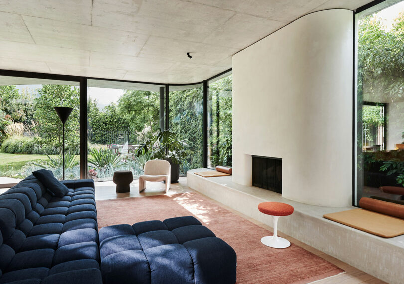 Modern living room with large windows, a navy sectional sofa, minimalist fireplace, indoor plants, and views of a lush garden outside.