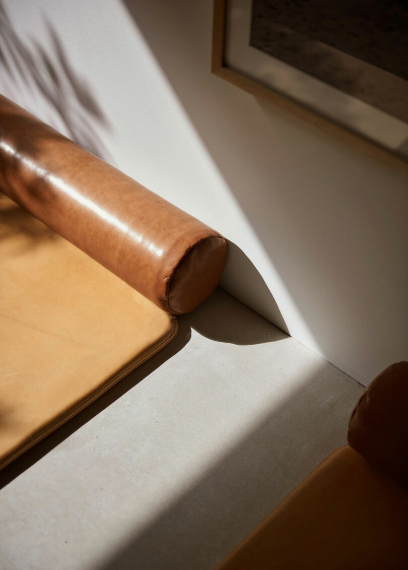 A close-up of a tan leather bench with cylindrical armrests, next to a white wall and a framed picture, in natural sunlight.