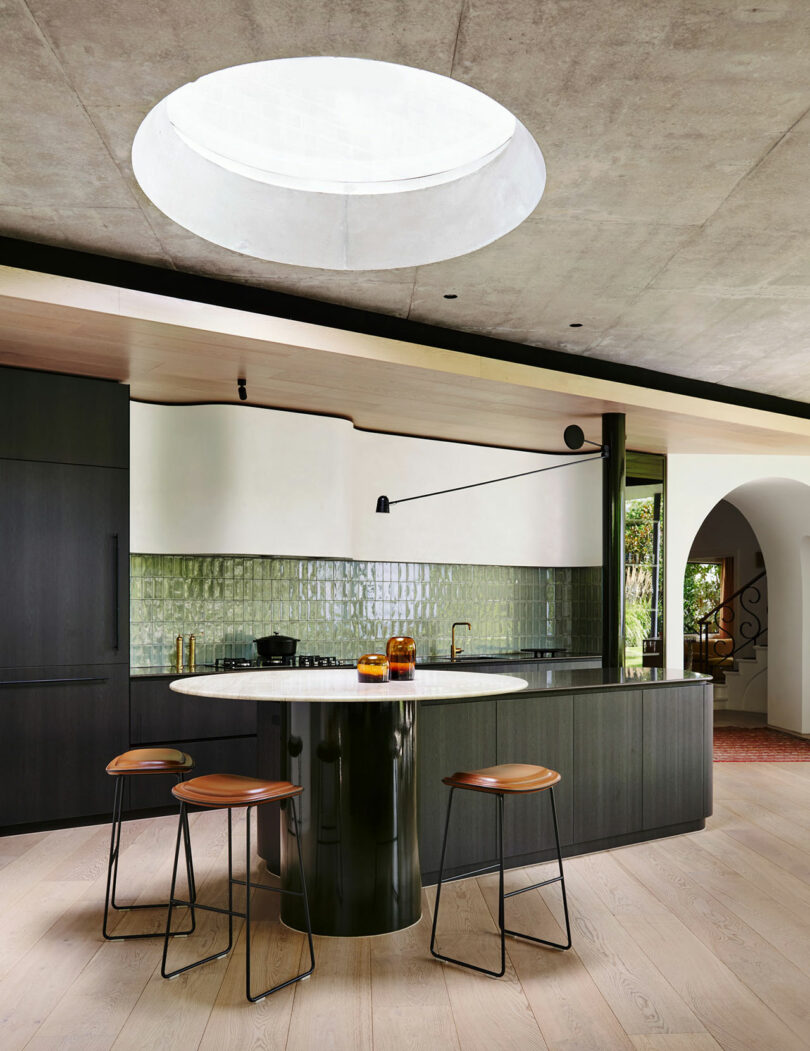 Modern kitchen with a round skylight, dark cabinetry, green tile backsplash, a curved island with a marble top, and three wooden bar stools on light wood flooring.