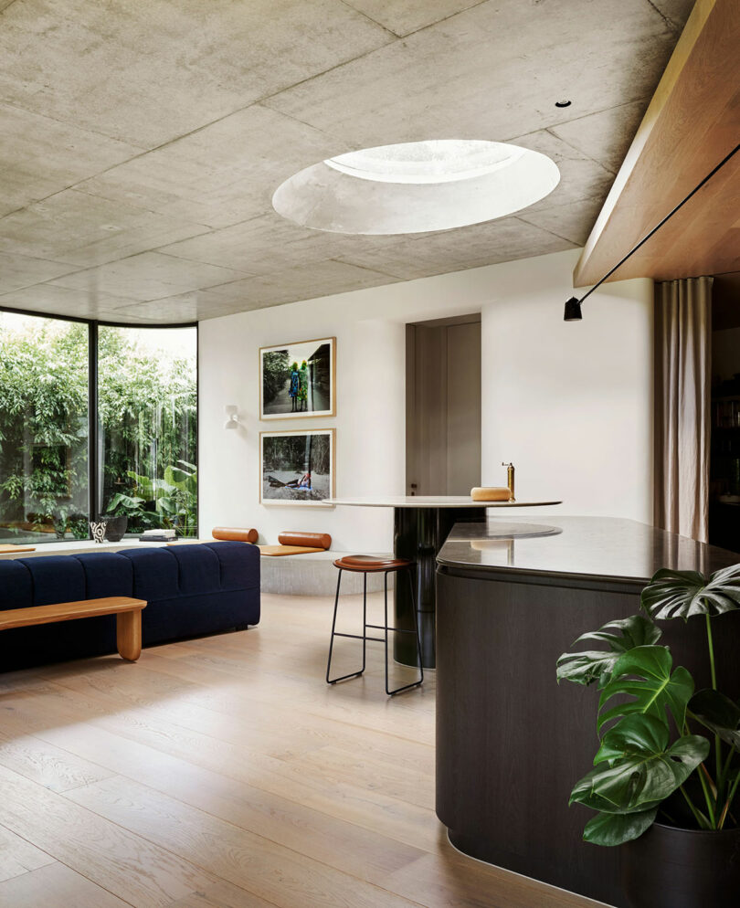 Modern living space with a concrete ceiling, circular skylight, dark sofa, kitchen island with stools, artwork on the wall, and large windows overlooking greenery.