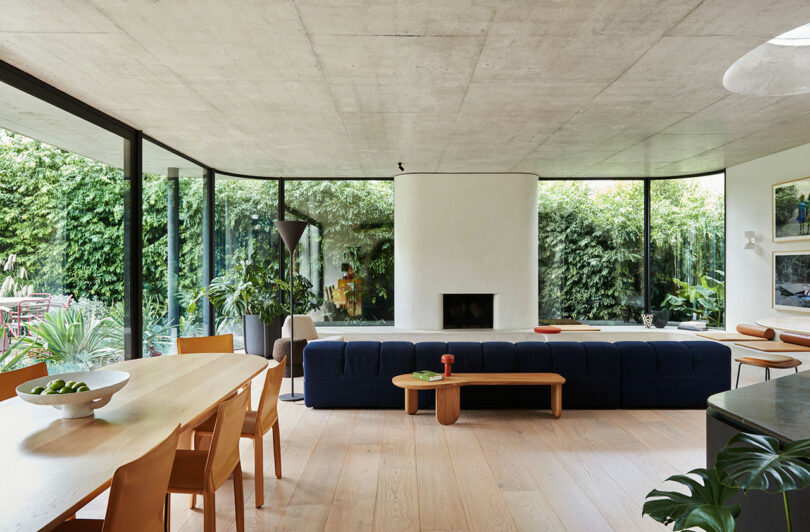 Modern living and dining area with large windows, a navy blue sofa, wooden furniture, indoor plants, and an outdoor view of greenery. A white fireplace is centered against a curved wall.