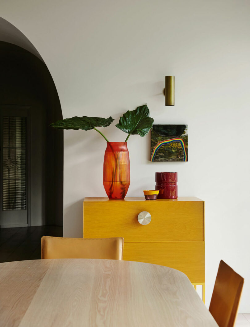 A yellow cabinet with decorative vases and a plant stands against a white wall, near a round wooden table and an arched doorway.