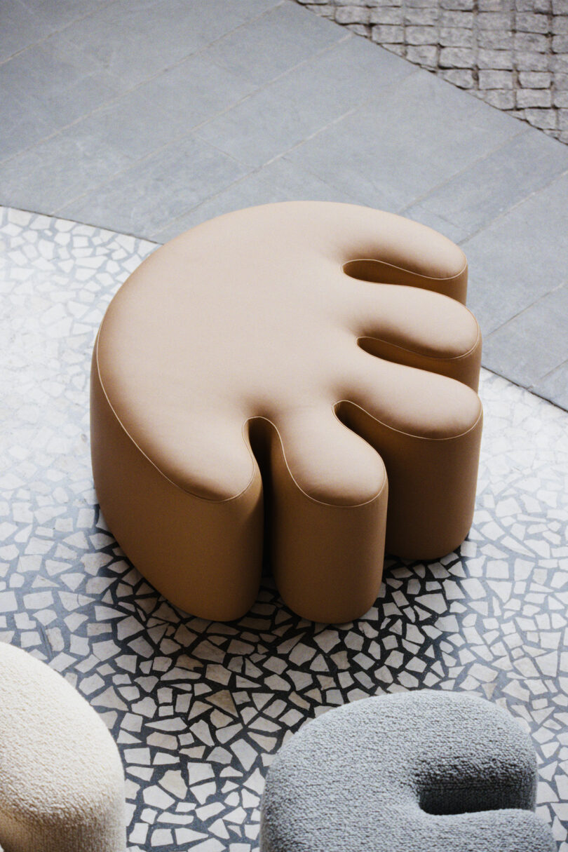 Beige, abstract-shaped ottoman with rounded, finger-like protrusions on a mosaic tile floor, next to similarly shaped furniture pieces.