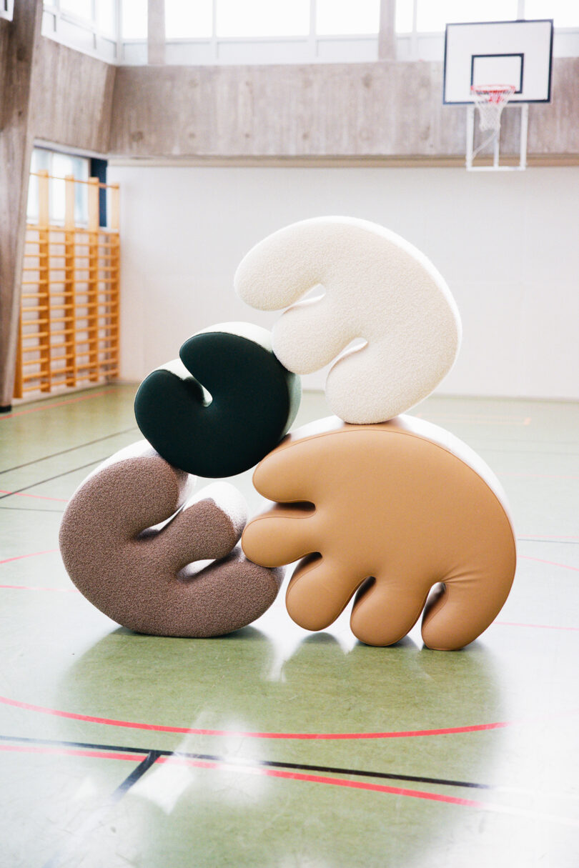 Four abstract, hand-shaped cushions in different colors are stacked on top of each other in the middle of an indoor gymnasium with a basketball hoop.