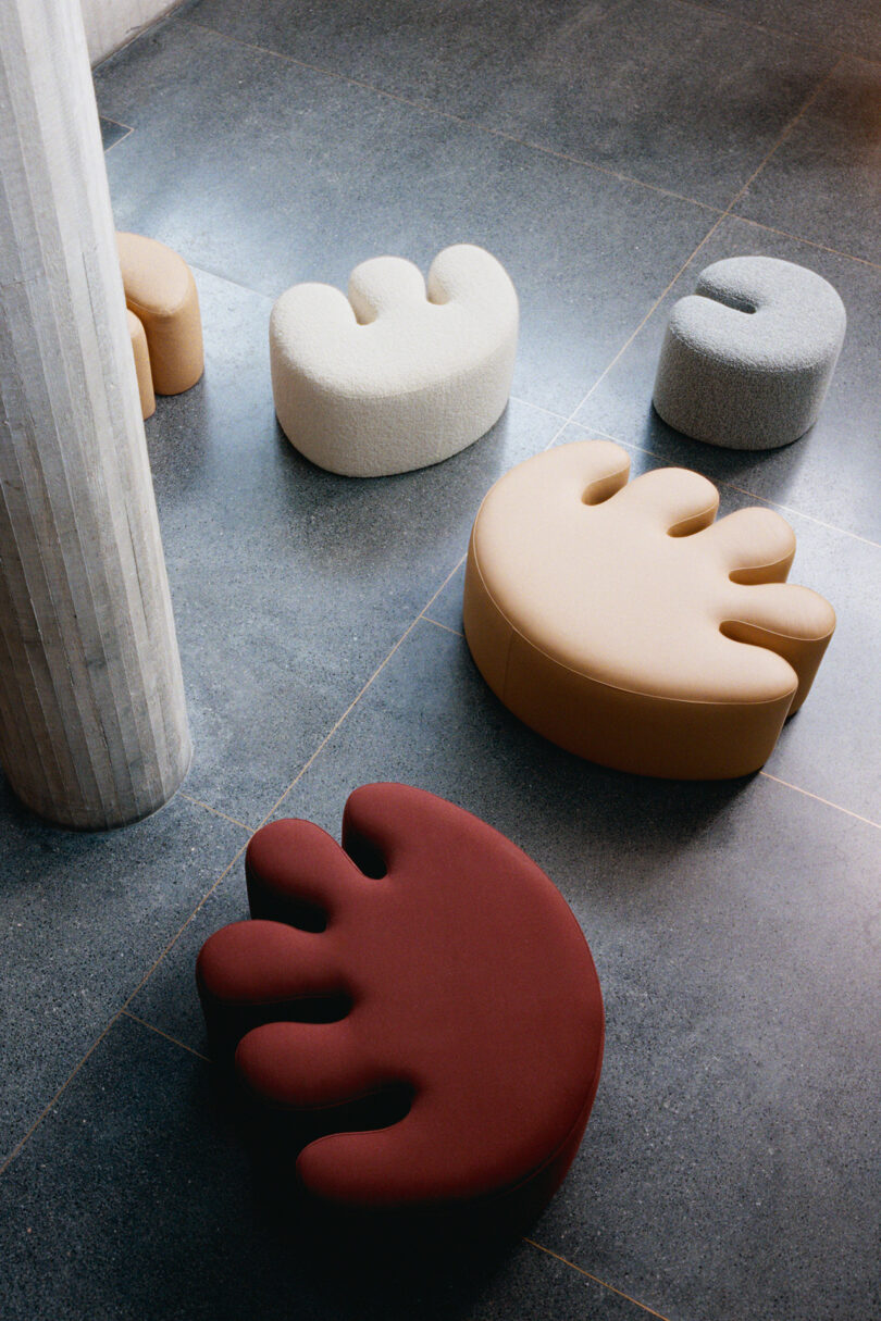 Five abstract, hand-shaped seats in different colors—cream, gray, beige, and red—are arranged on a dark stone floor beside a tall, round column.