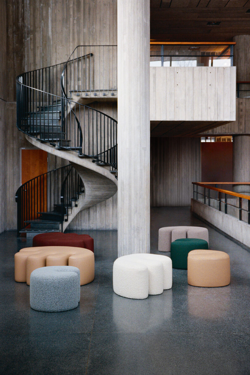 A modern interior with concrete walls, a black spiral staircase, and several round ottomans in neutral and green tones arranged on a polished floor.