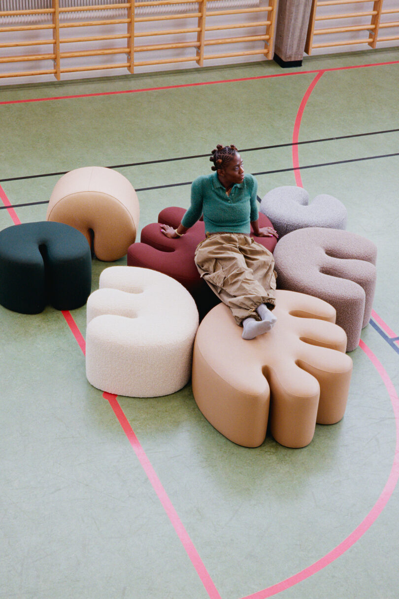 A person sits on modular, abstract-shaped poufs arranged in a circle on a gymnasium floor with red and black lines.
