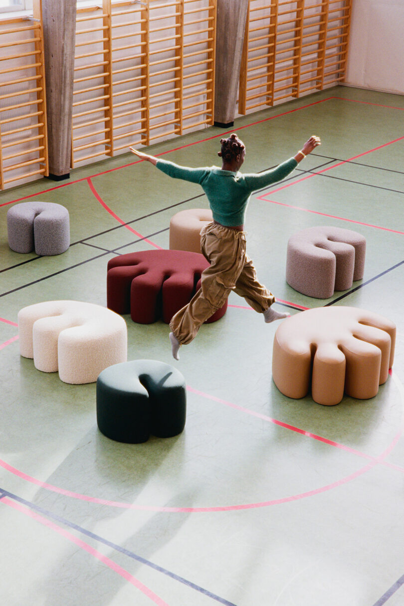 A person jumps across colorful, abstract-shaped stools arranged in a gymnasium with wooden wall bars in the background.
