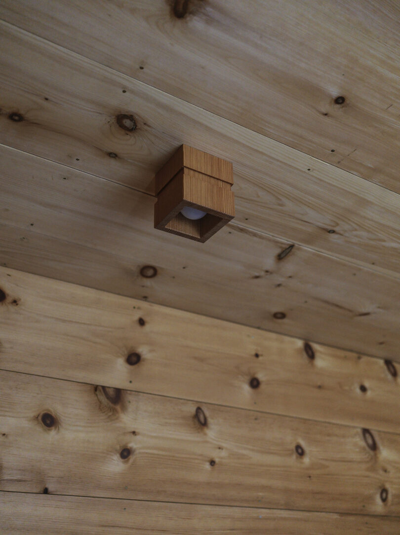 A small, square wooden light fixture is mounted on a wooden plank ceiling with a natural wood grain pattern.