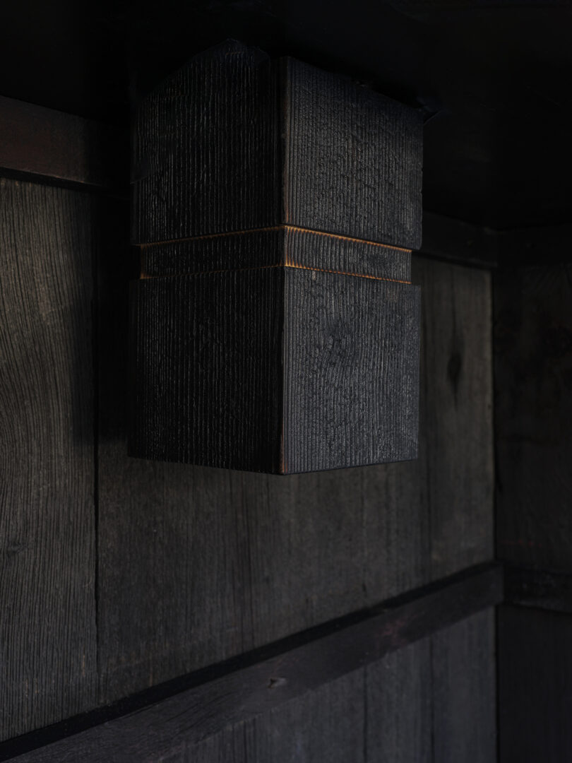 A close-up of a charred wooden structural beam with gold accent lines, set against a wall of dark, burnt wood panels.