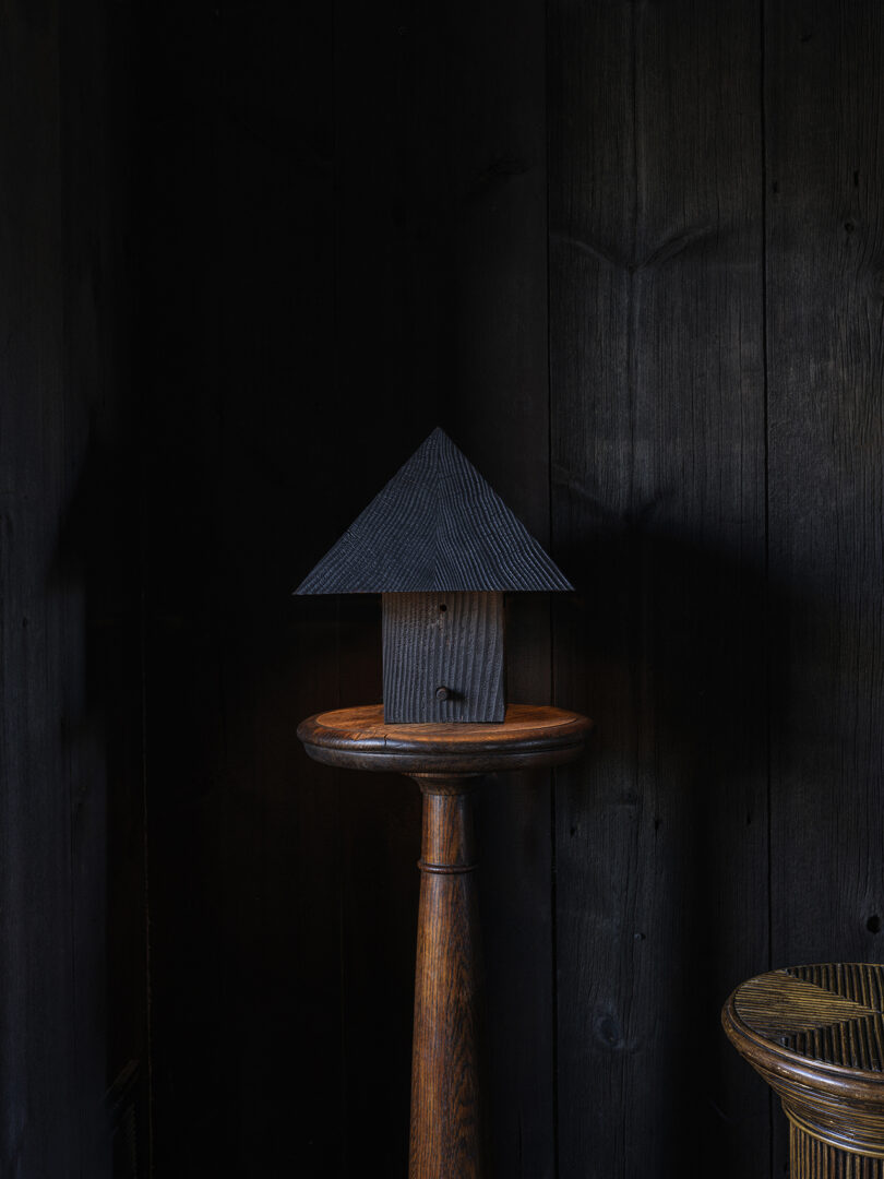 A small, dark, house-shaped object with a ridged roof sits on a wooden pedestal against a background of dark wooden panels.