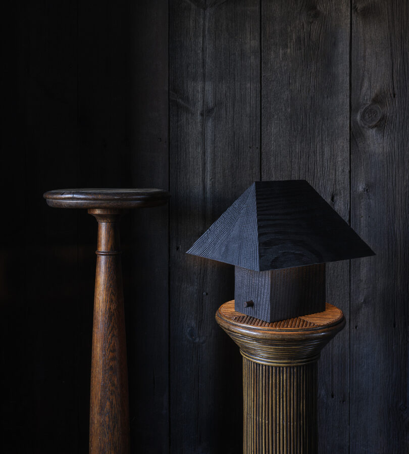Two wooden pedestals stand against a dark wood-paneled wall; one holds a small black house-shaped object with a pyramid roof.