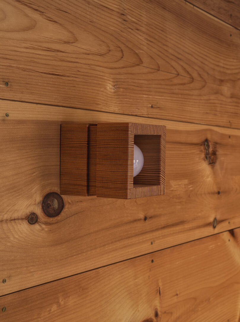 A wooden wall-mounted light fixture with a rectangular, slatted design exposes a white bulb, set against a wooden plank wall.