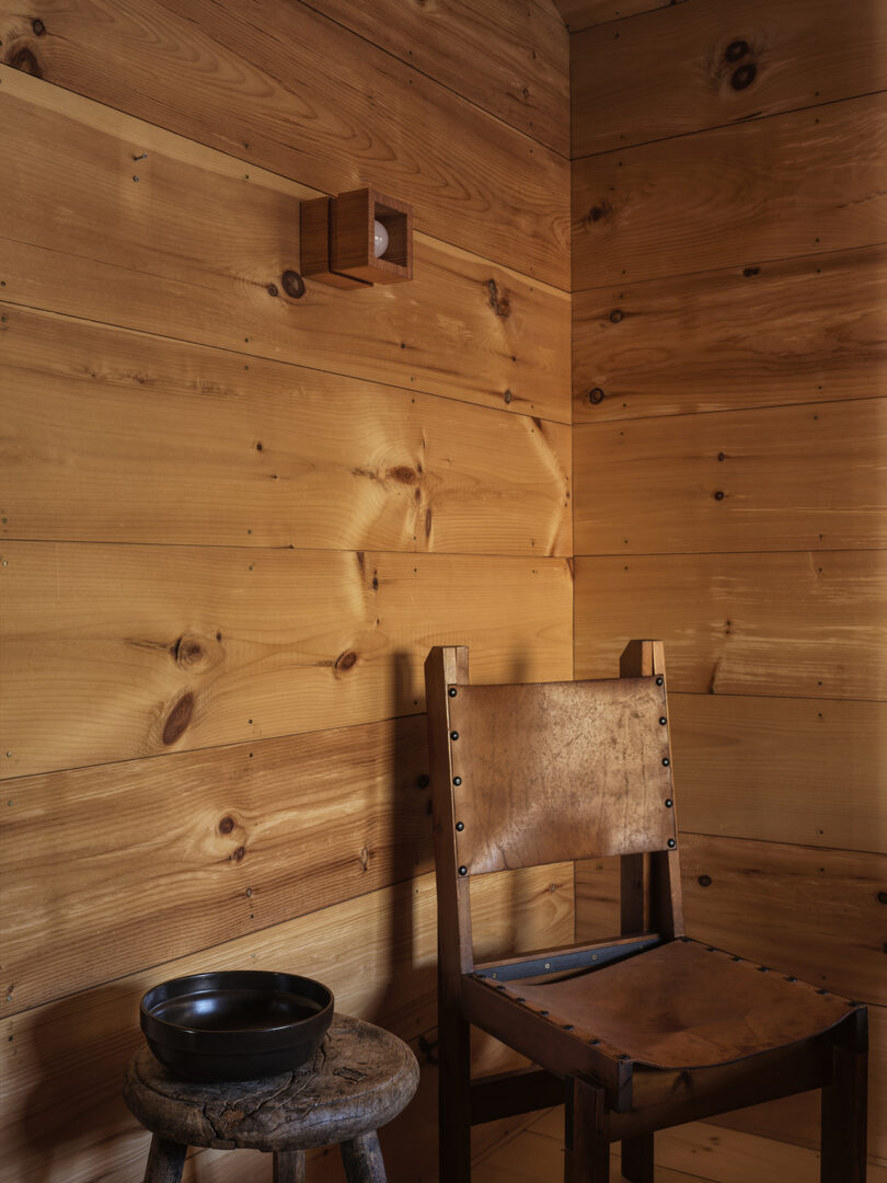 A wooden chair and small table with a black bowl are placed in the corner of a room with wood-paneled walls and a small modern wall light.