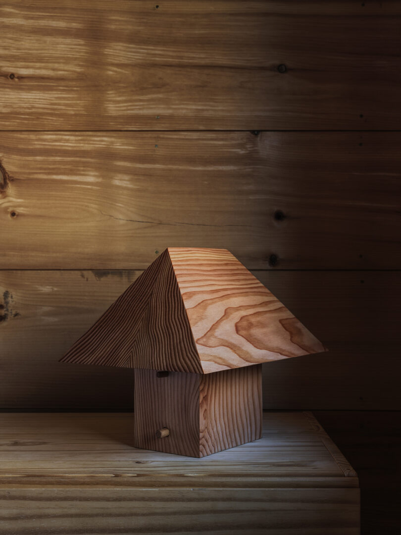 A small wooden structure with a square base and a large, slanted pyramid-shaped roof sits on a wooden surface against a wooden wall.