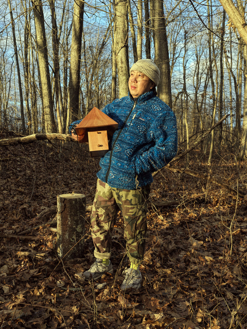 A person wearing a blue jacket, camouflage pants, and a knit hat stands in a forest holding a wooden birdhouse.