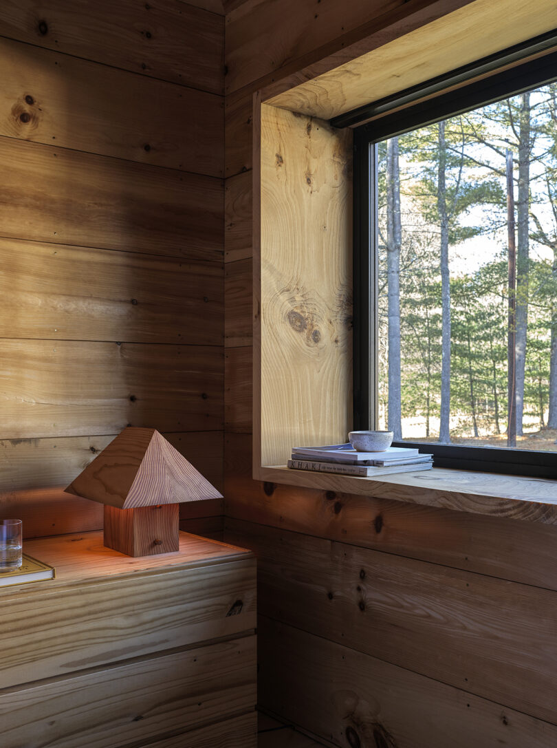 A wooden interior features a pyramid-shaped lamp, a glass of water on a table, and a window ledge with stacked books and a cup, overlooking trees outside.