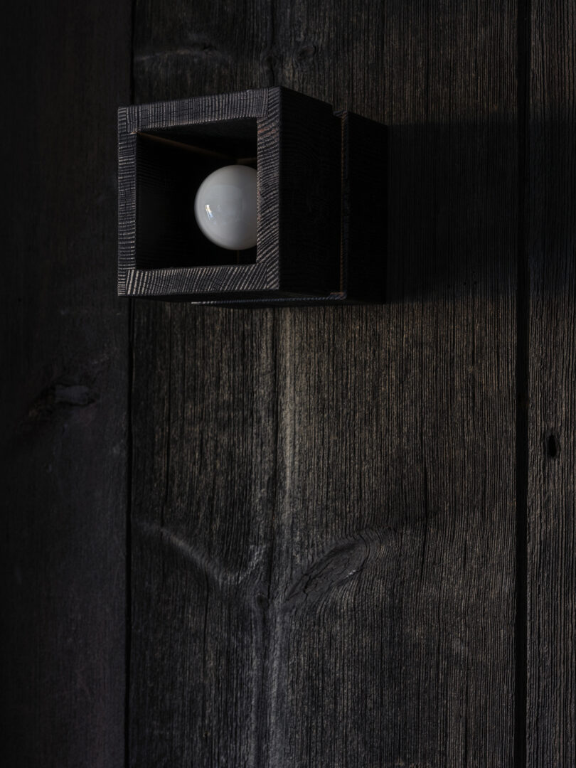 A square wooden wall sconce with a visible light bulb is mounted on a dark, textured wooden wall.