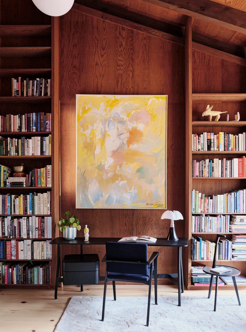 A modern home office with wooden shelves filled with books, a large abstract painting on the wall, a black desk, a chair, and a table lamp on a light rug.
