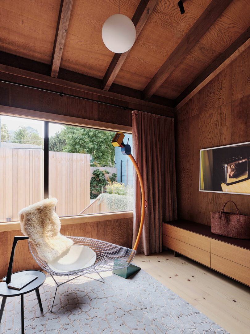 A modern room with wood paneling, a large window, a wire lounge chair with a fur throw, an orange floor lamp, a wall-mounted TV, a round side table, and a rug.