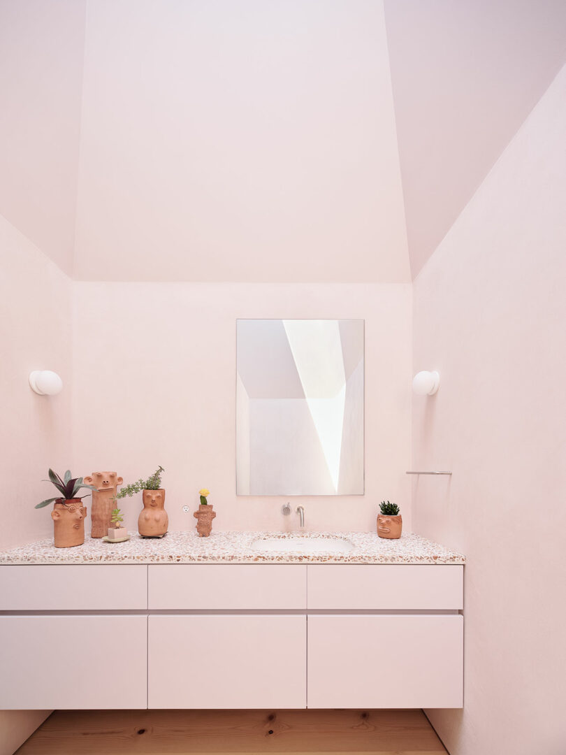 A minimalist bathroom with a white vanity, terrazzo countertop, rectangular mirror, and potted plants in clay pots against light pink walls.