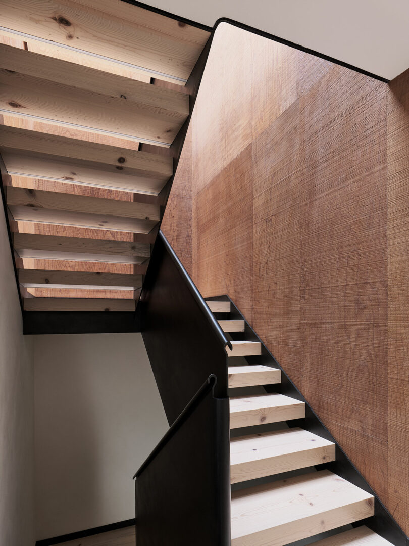 Modern wooden staircase with open risers, black metal railing, and a textured wooden wall panel alongside the stairs.