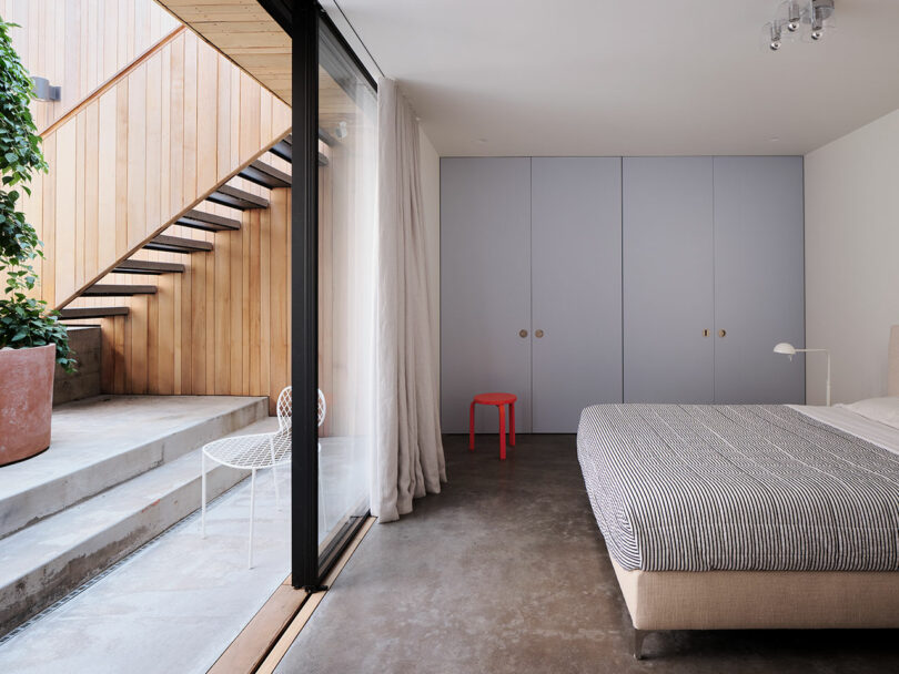 Modern bedroom with a bed, gray closet, small red stool, and sliding glass door opening to a patio with wooden stairs and potted plant.