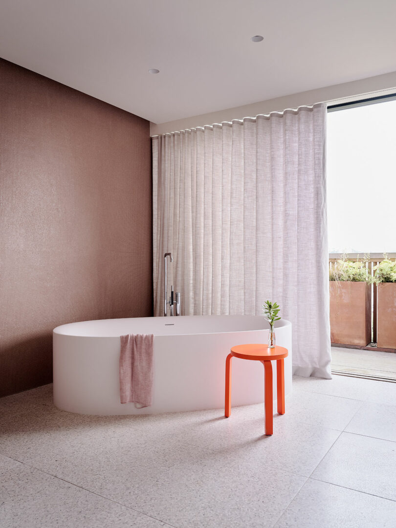 A modern bathroom with a white oval bathtub, a pink towel, a small orange side table with a plant, sheer curtains, and an open sliding door to a terrace.