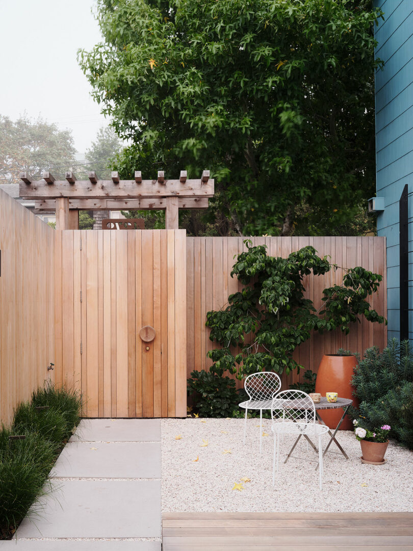 A small backyard patio with a wooden fence, a metal table and two chairs, potted plants, and trees in the background.