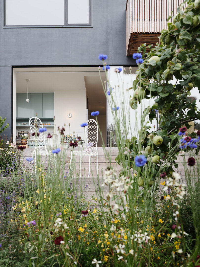 Wildflowers grow in the foreground of a garden, with a modern house, patio, and white chairs visible through open glass doors in the background.