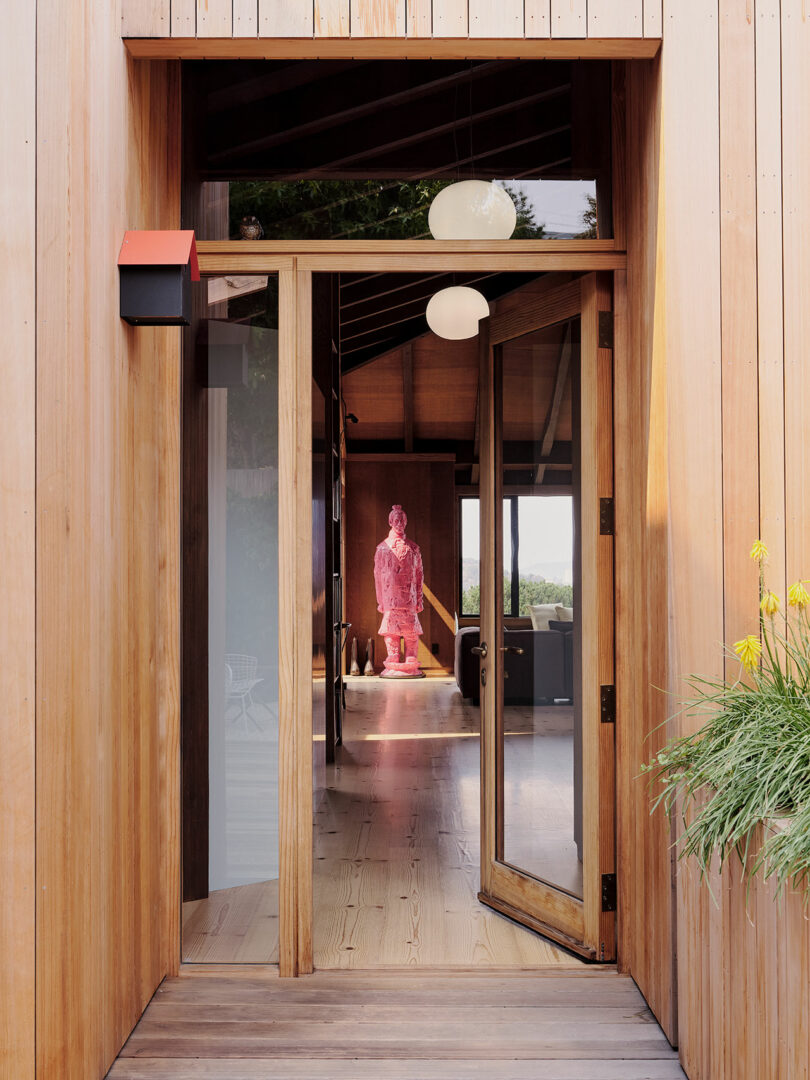 Open wooden door reveals modern interior with a pink statue in the center, hanging round lights, and potted yellow flowers at the entrance.